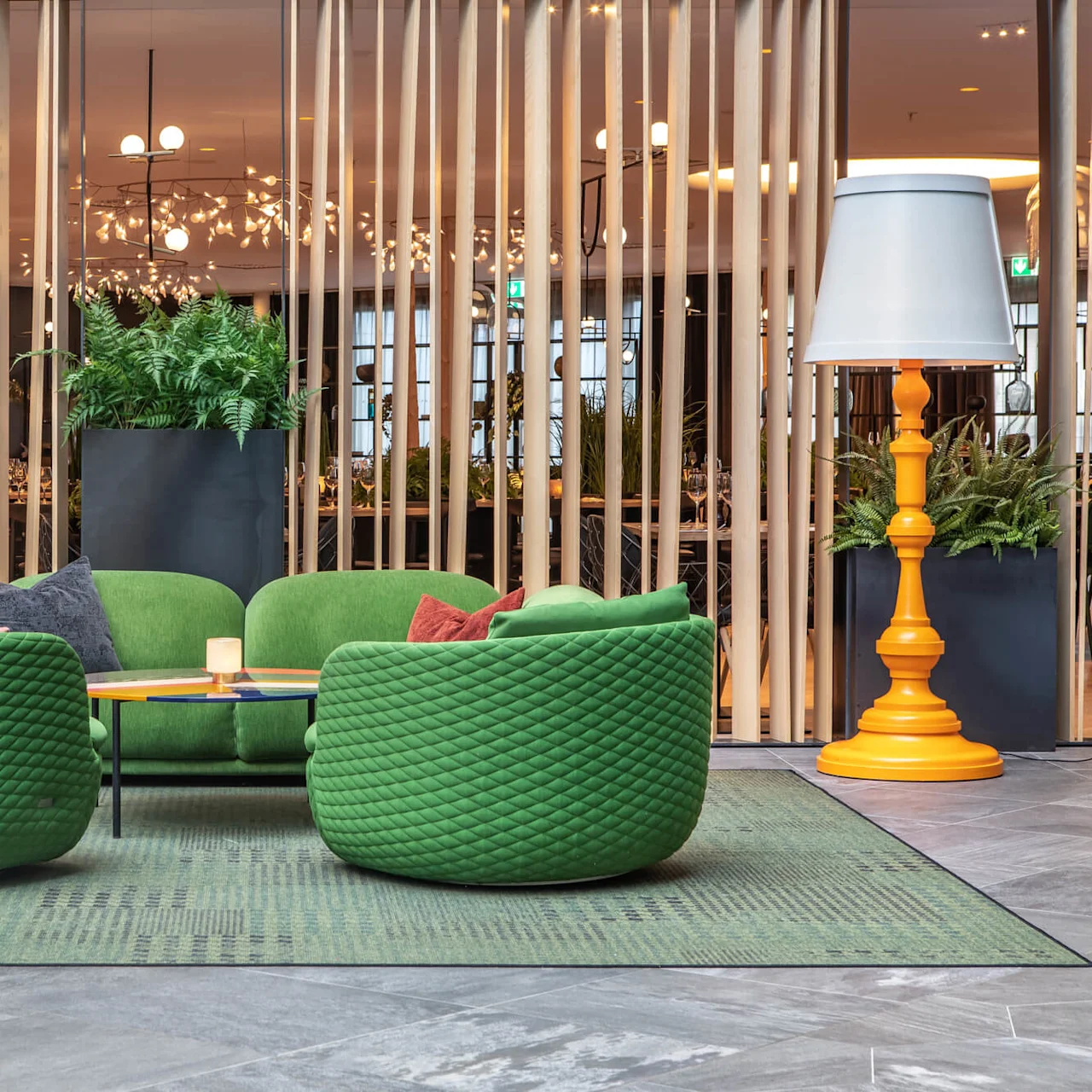 Four green, tufted armchairs encircle a low table with a candle on it, surrounded by a modern lobby with wooden dividers and potted plants.