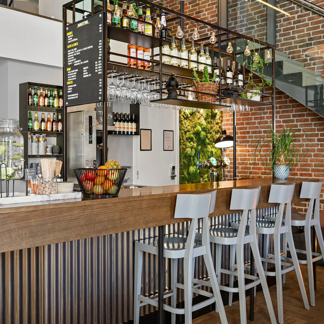 Bar counter with high chairs, displaying bottles and glasses on shelves. Menu board lists: Snacks, Bites, Beer & Cider, Wine, Beverages, Snacks, and Sweets. Brick wall and indoor plants surround. 