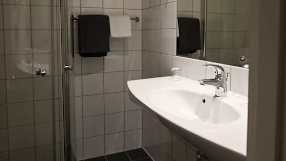 Ett handfat med kromkran är monterat på en kaklad vägg, med handdukar hängande bredvid och ett glas på bänken i ett modernt badrum.