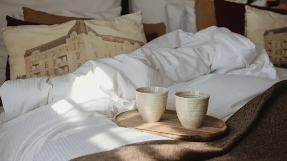 Deluxe-hotellihuoneen sänky raikkailla valkoisilla lakanoilla, ainutlaatuisella tyynyllä ja kahdella lämpimällä juomalla tarjottimella, aamuauringon valossa.