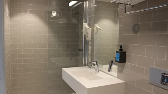 A modern bathroom shows a white sink with a mounted hairdryer, surrounded by beige tiles. Above, a towel rack with folded towels. To the right, a hand wash dispenser is attached.