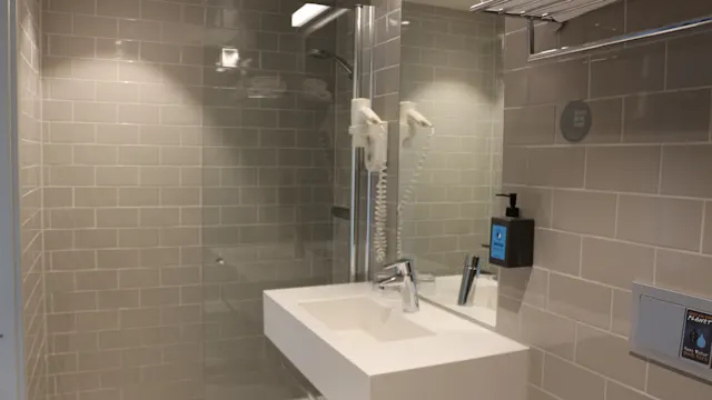 Ett modernt badrum visar ett vitt handfat med monterad hårtork, omgiven av beige kakel. Ovanför finns en handdukshylla med vikta handdukar. Till höger är en tvåldispenser monterad.