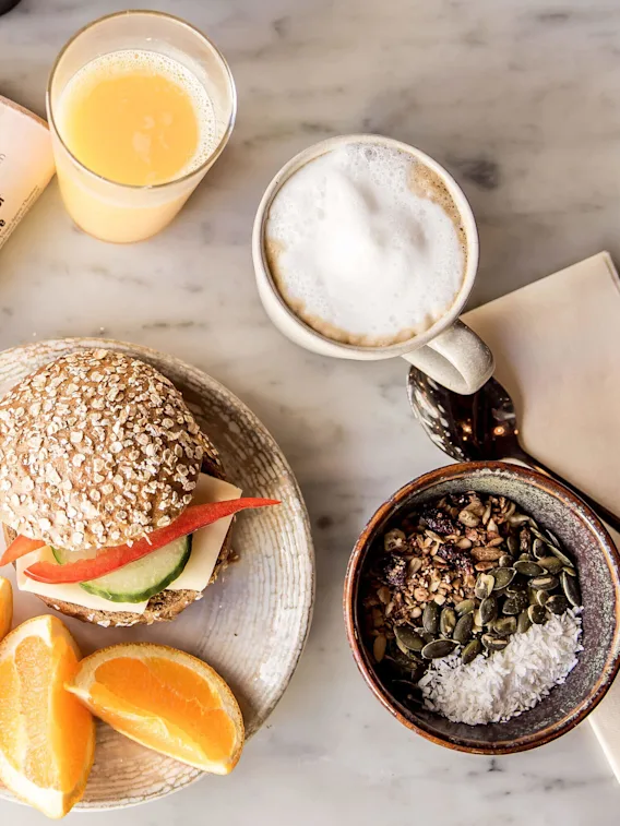 En morgenmadsspredning på et marmorbord med en sandwich, appelsinskiver, kaffe, juice, og granola, ledsaget af læsebriller på en avis.