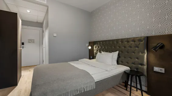 Ett modernt hotellrum med en stor säng, grå och vita sängkläder och mönstrad tapet, redo för en gäst.