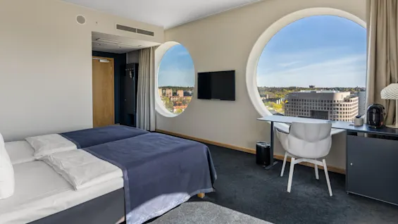Et moderne hotelværelse med to senge, et skrivebord og store cirkulære vinduer med en lys byudsigt.