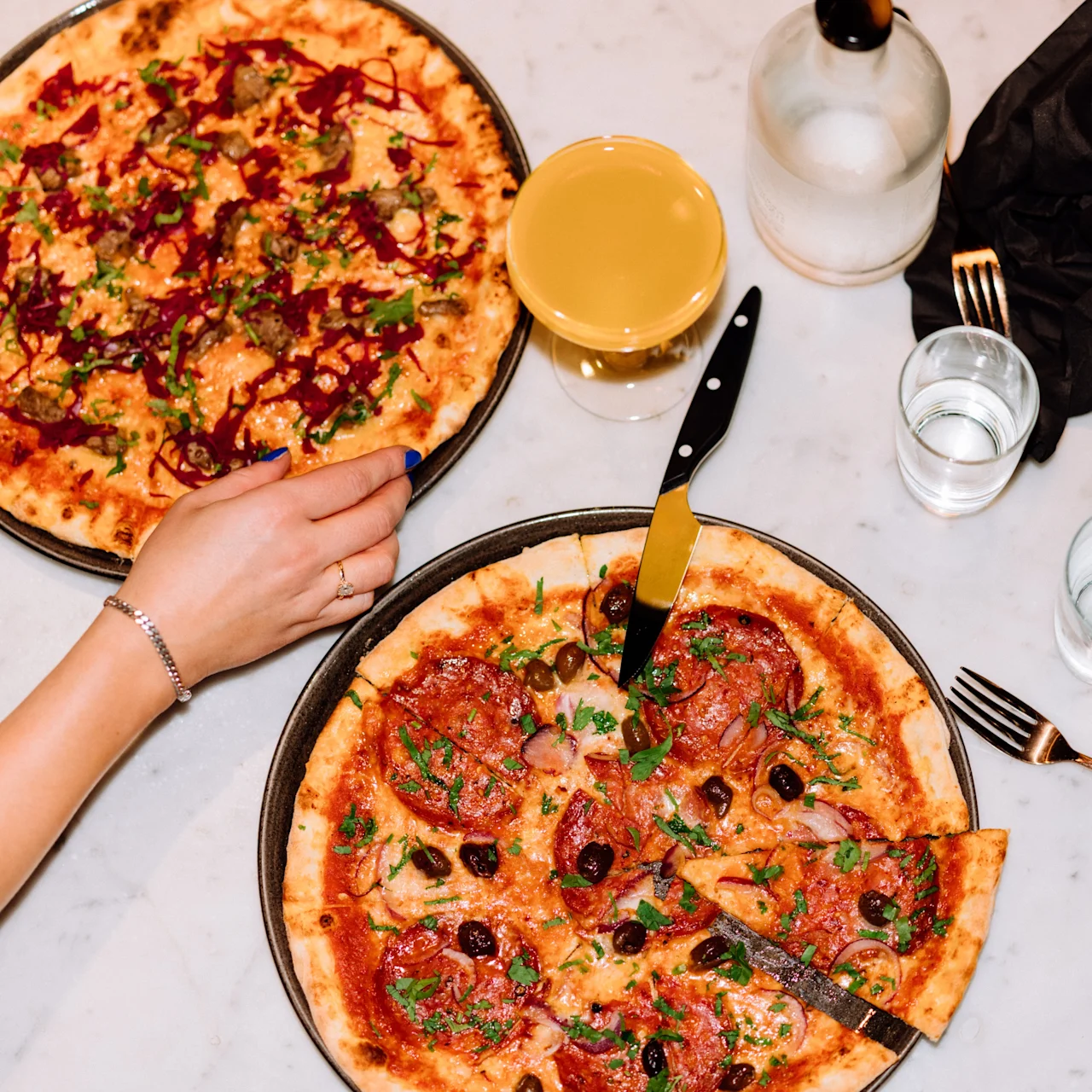 Two pizzas, one with sliced meat and the other with vegetables, placed on a marble table. A hand reaches for a slice. Nearby are glasses, utensils, and a bottle.