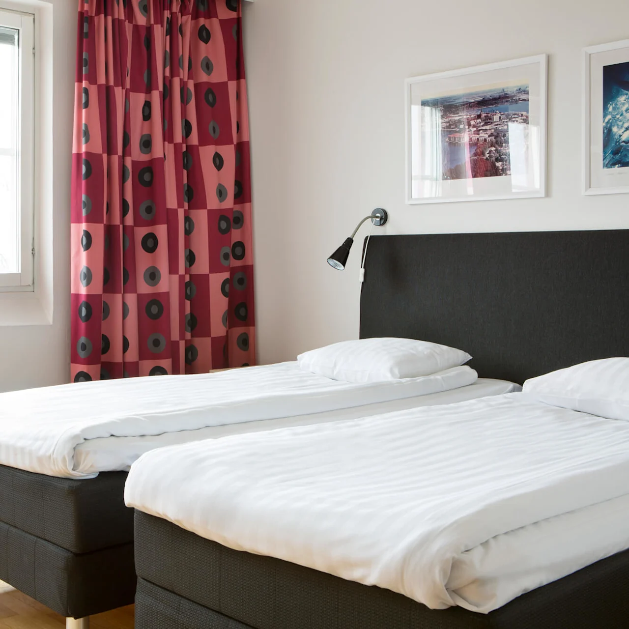 Two neatly made twin beds sit side by side in a bright room with red patterned curtains, white walls, and framed artwork. A modern chair is nearby.