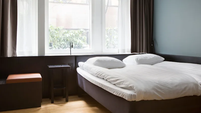 Bed with white linens is neatly arranged, positioned beside large windows with white curtains, sunlight streaming into the modern, minimalist room.