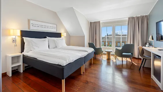 A bright, modern hotel room with a large bed, seating area, and desk, illuminated by natural light from large windows.