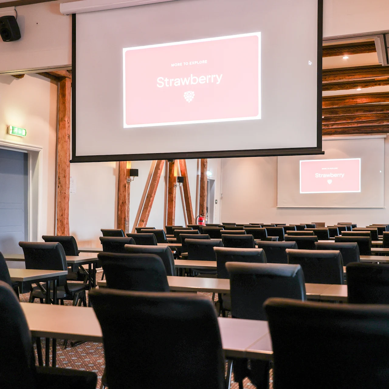 Projector screen displays "Strawberry" in a conference room, featuring rows of empty chairs facing the screen. Wooden beams and a small wall clock add to the room's design.