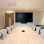 A bright and modern conference room with a long wooden table, comfortable seating, a large screen for presentations, and a whiteboard. Exit.
