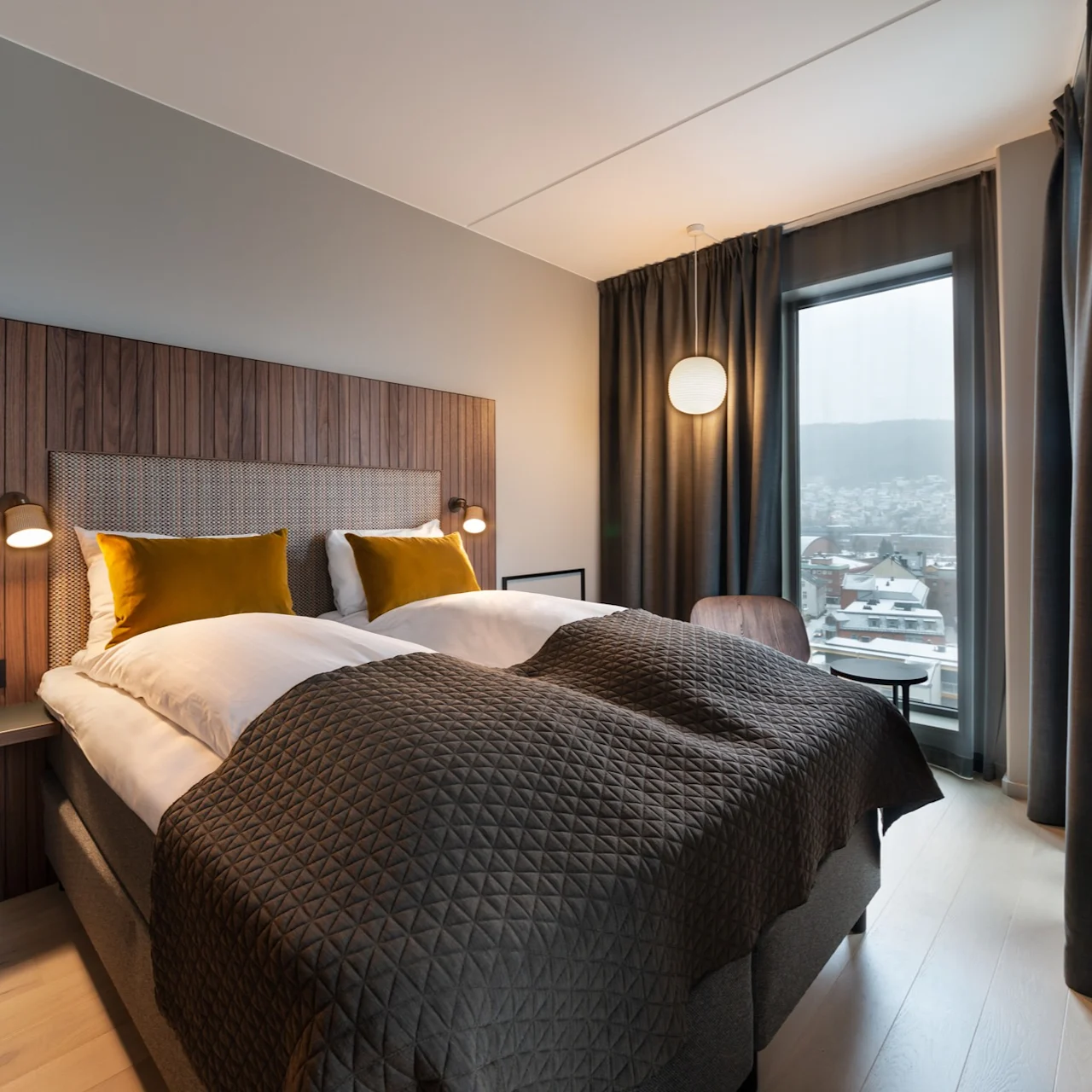 Et moderne hotellrom har en dobbeltseng med sennepsgule puter og et brunt teppe, omgitt av trepaneler. Et vindu tilbyr utsikt over snødekte hustak i en urbant landskap.