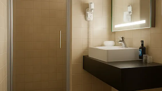 Et moderne bad med beige fliser, et stilig vask, et stort speil med lys og en dusj. En hårføner henger på veggen, og noen nødvendigheter er pent plassert ved vasken.