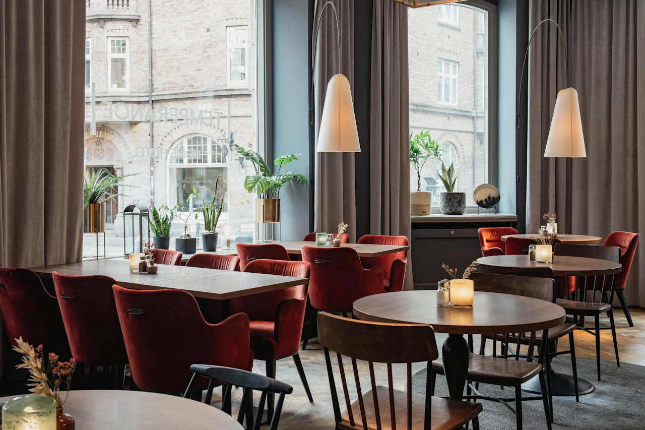 Innbydende restaurantinteriør med røde stoler, trebord og myk belysning. Store vinduer avslører en bygata og TEMPEL HOTEL.