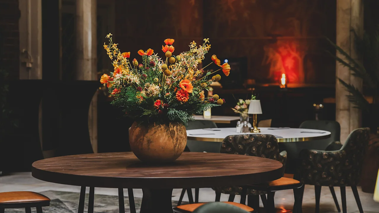 A large bouquet of vibrant flowers sits in a round vase on a wooden table, surrounded by cushioned chairs in a softly lit, elegant dining area.