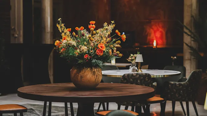 En stor bukett med fargerike blomster står i en rund vase på et trebord, omgitt av polstrede stoler i et mykt opplyst, elegant spiseområde.