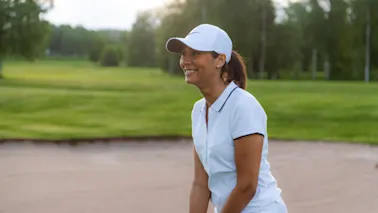 En person i hvit caps og pikétrøye smiler mens de nyter en golfrunde på en solrik dag.