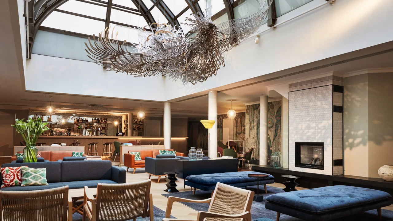 An intricate chandelier hangs in a modern lounge with sofas, armchairs, and a fireplace under a glass dome ceiling.