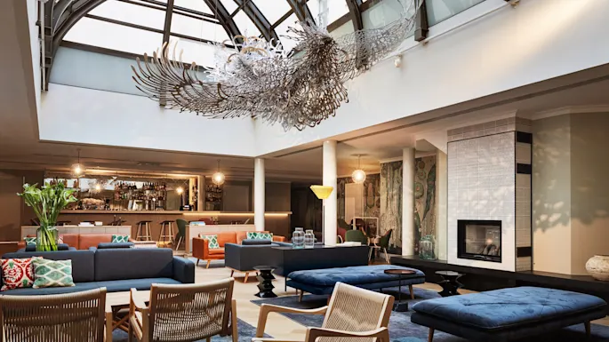 En intrikat lysekrone henger i en moderne salong med sofaer, lenestoler og en peis under et glasskuppeltak.