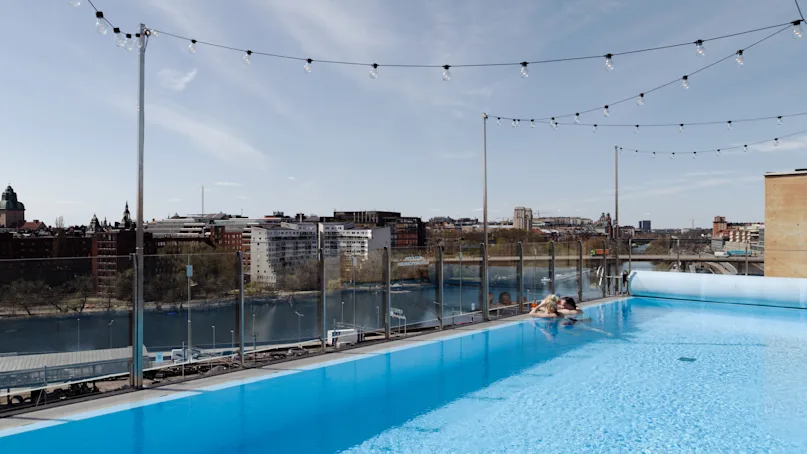 Rooftop pool with a person relaxing at the edge, surrounded by string lights. Overlooks a cityscape with buildings and a river under a clear sky.