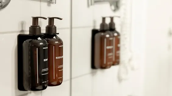 Två pumpflaskor märkta SOAP och BODY LOTION är monterade på en vit kaklad vägg i en badrumsmiljö.