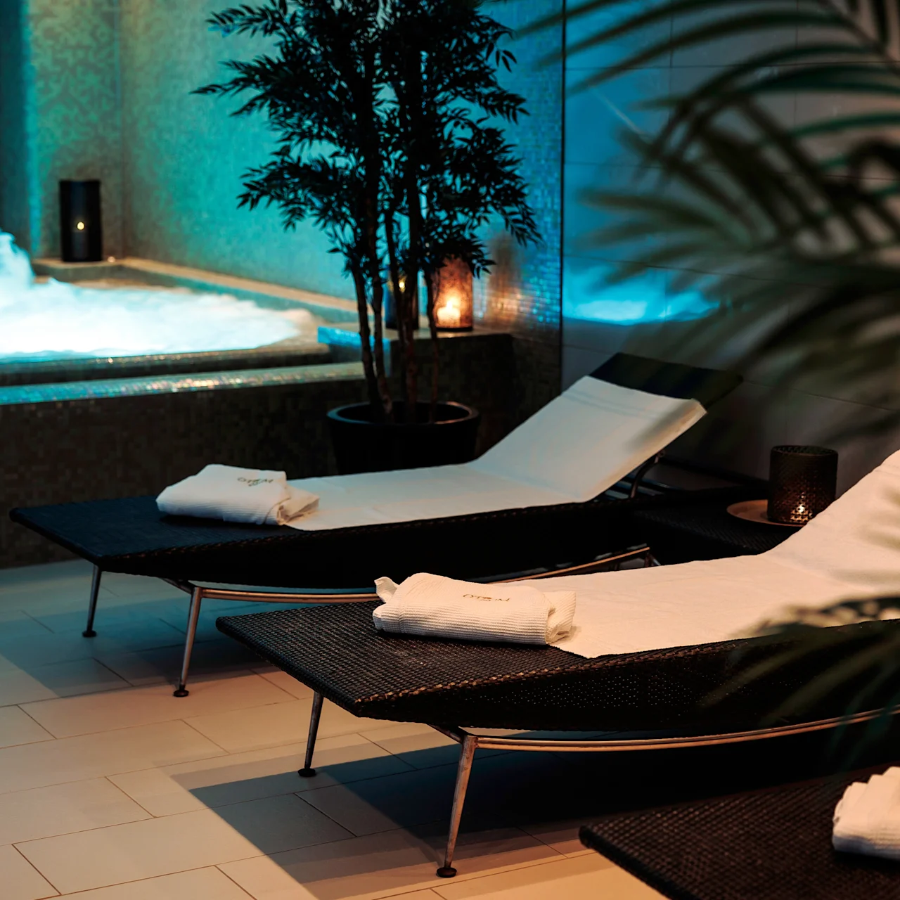 Loungers are aligned with towels on them, next to a bubbling hot tub. Ambient lighting and potted plants create a calming spa atmosphere.
