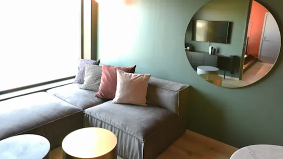 En grå L-formet sofa med farverige puder er placeret ved et stort vindue. Et rundt spejl reflekterer et væghængt TV og en gang i en moderne stue.