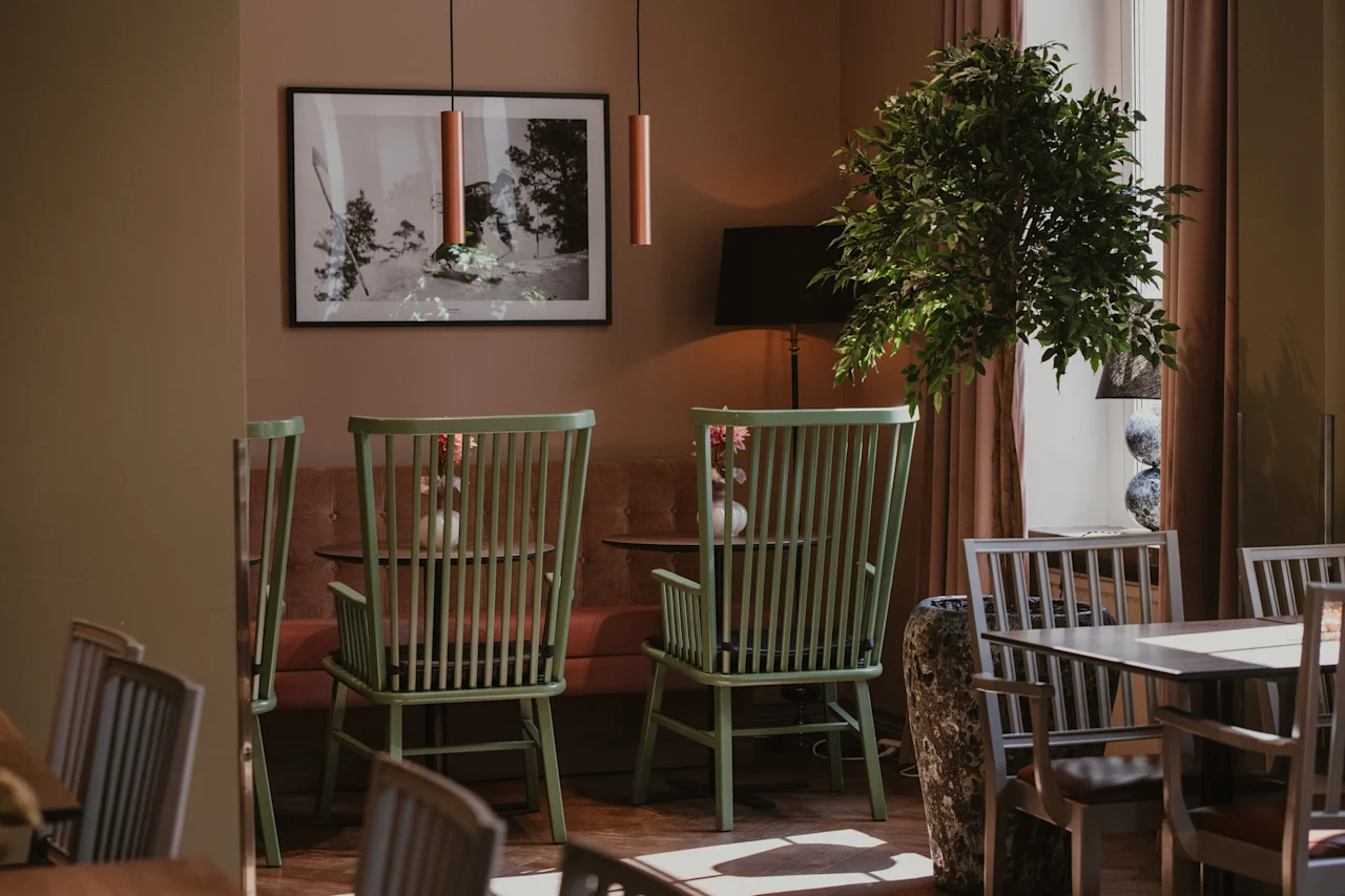 Et varmt og innbydende spiseområde med lysegrønne stoler, en stor plante og et innrammet kunstverk på veggen, badet i naturlig lys.
