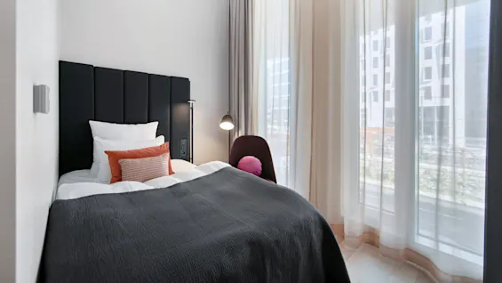 A bed with dark headboard and gray blanket sits beside a window with sheer curtains. An orange and white pillow add contrast, while a floor lamp stands nearby.