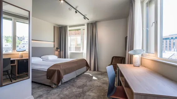 En pænt redt seng med brune og hvide lagener står ved store vinduer i et moderne, lyset hotelværelse. Et skrivebord med stol og lampe er i nærheden.