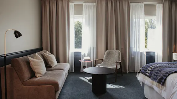 Ett bekvämt rum med en soffa, en fåtölj och en säng, upplyst av solljus som strömmar in genom draperade fönster.