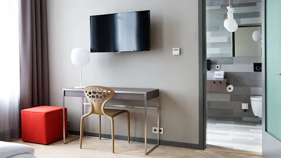 Et moderne hotellrom har en veggmontert TV, et skrivebord i tre med en unik stol og en rød puff. En åpen dør avslører et stilig bad med toalett og håndklestativ.