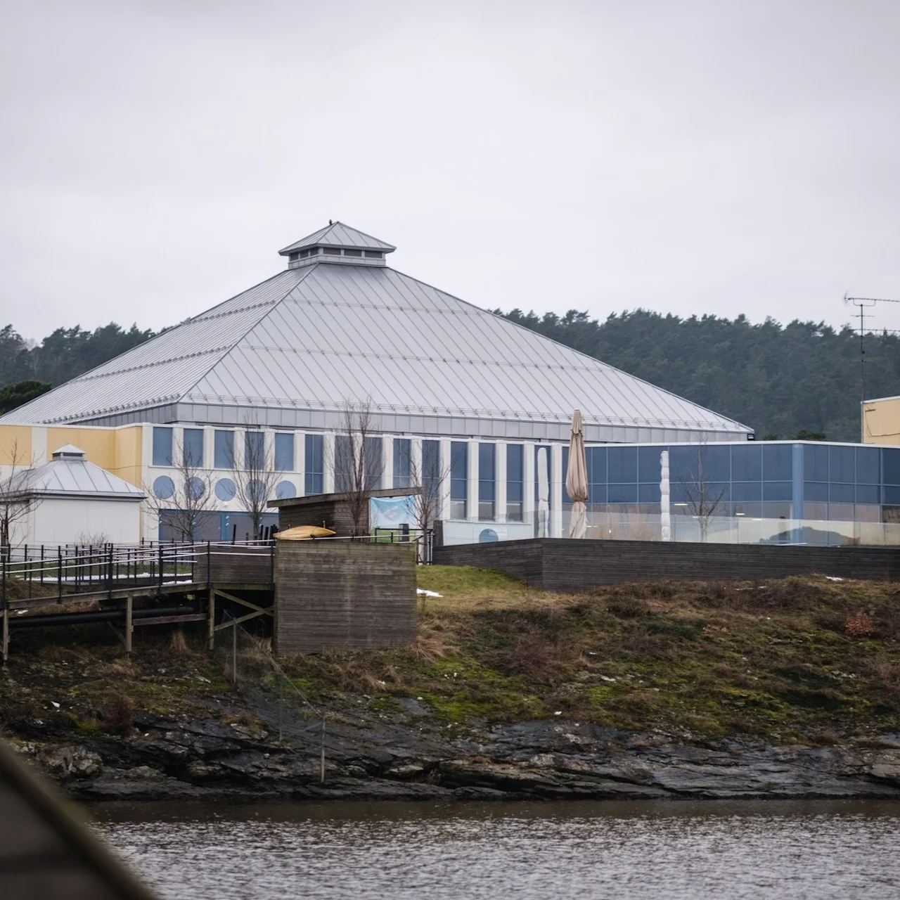 A large, pyramid-shaped building with glass walls stands on a grassy hill by a river, surrounded by a forest in the background under an overcast sky.
