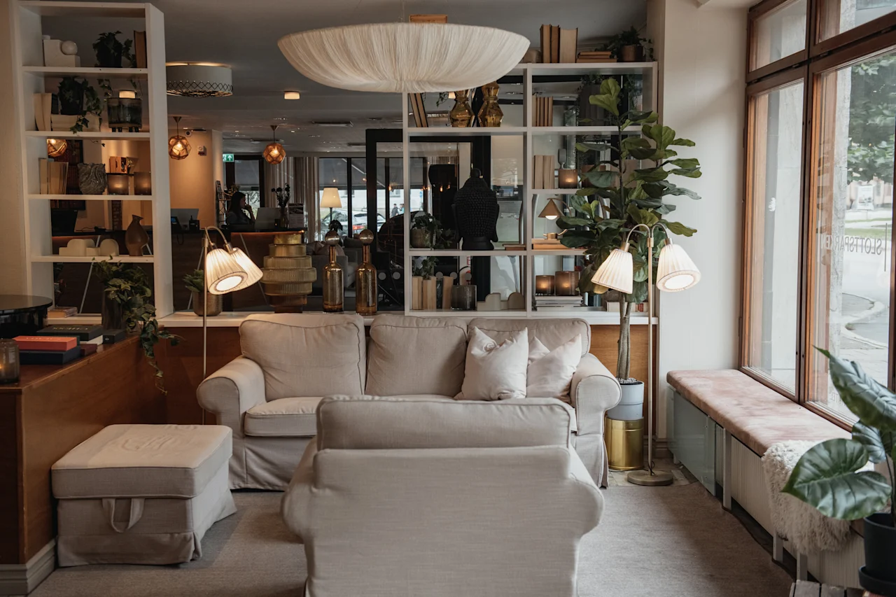 A cozy hotel lounge with comfortable sofas, a large bookshelf, and a window seat, creating a welcoming atmosphere. Visible text: HOTEL