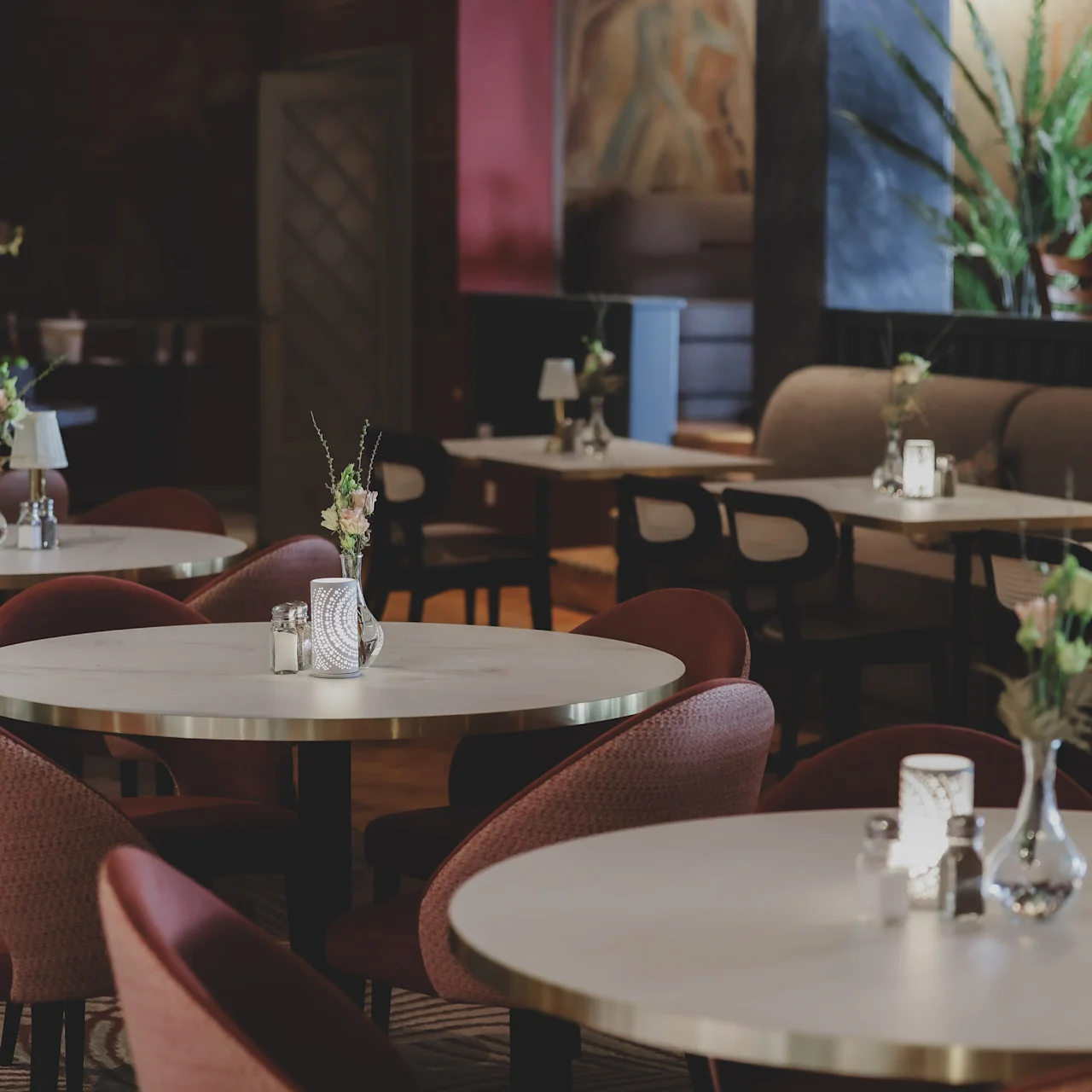Tables are set with small vases and candles in a dimly lit, elegant restaurant with red chairs and potted plants. The decor includes artwork on the walls, creating a sophisticated atmosphere.