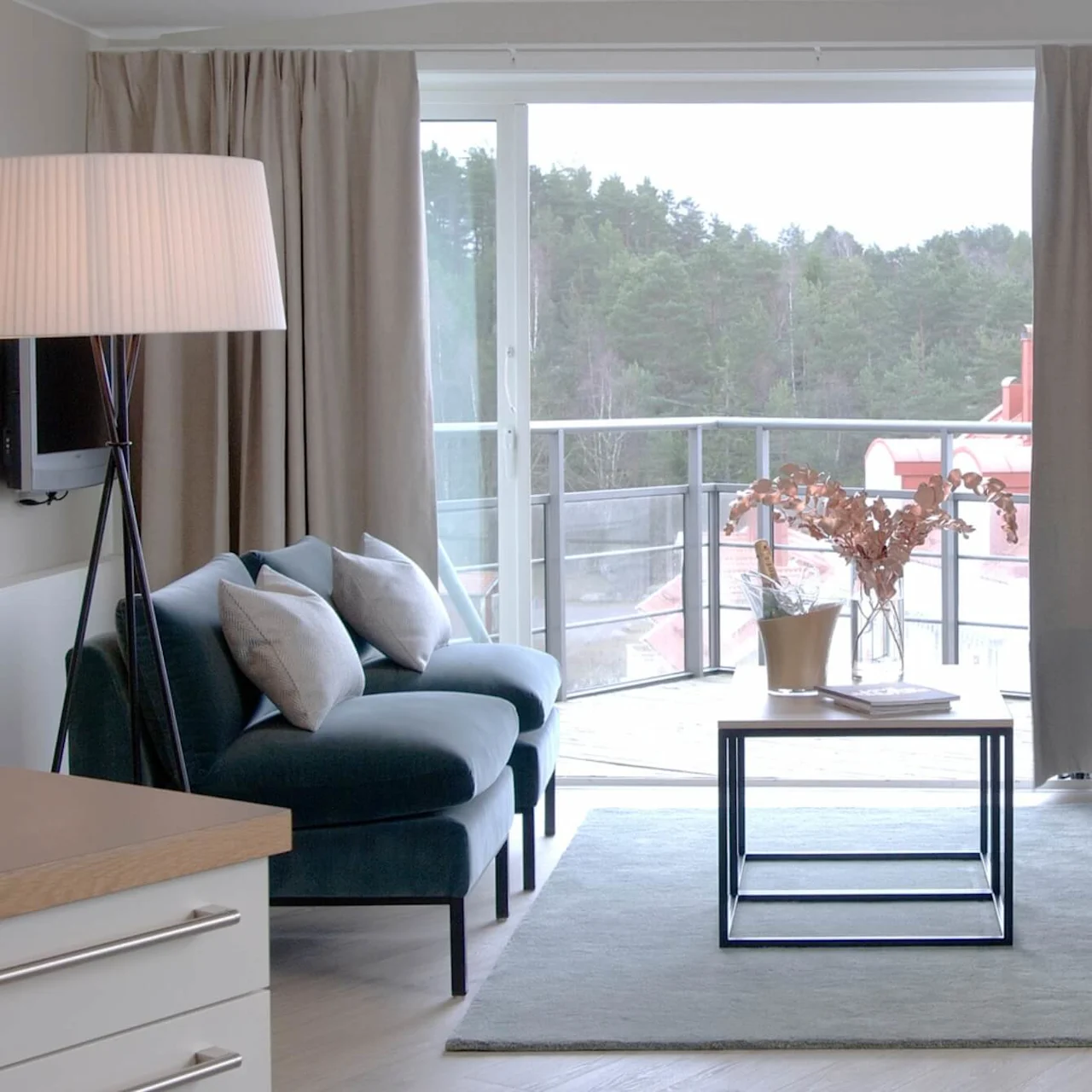 En moderne stue med en beige sofa og en blå lenestol rundt et salongbord med tørkede blomster på et teppe. Store vinduer viser en balkongutsikt mot skogen.