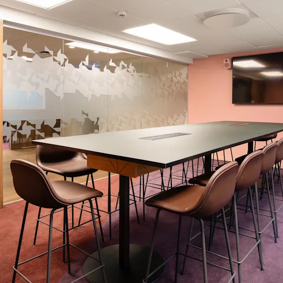 A long black table with brown chairs encircles a modern conference room. Frosted glass walls feature abstract patterns, and a large screen is mounted on a light peach wall. Text: None.