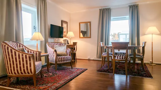 A room with striped armchairs and a wooden dining table, surrounded by curtains and lamps, features a wall-mounted TV and patterned rugs on a wooden floor.