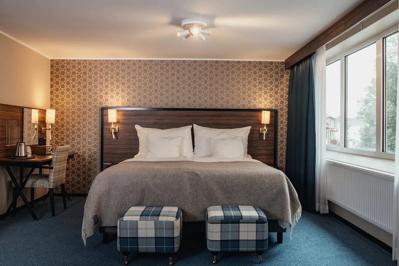 A welcoming hotel room featuring a comfortable bed, stylish plaid ottomans, and a bright window.