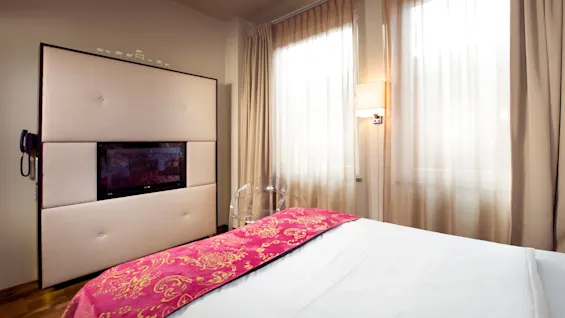 En pent oppredd seng med rødt og gullfarget overkast i et rom med gjennomsiktige gardiner og en veggmontert TV.
