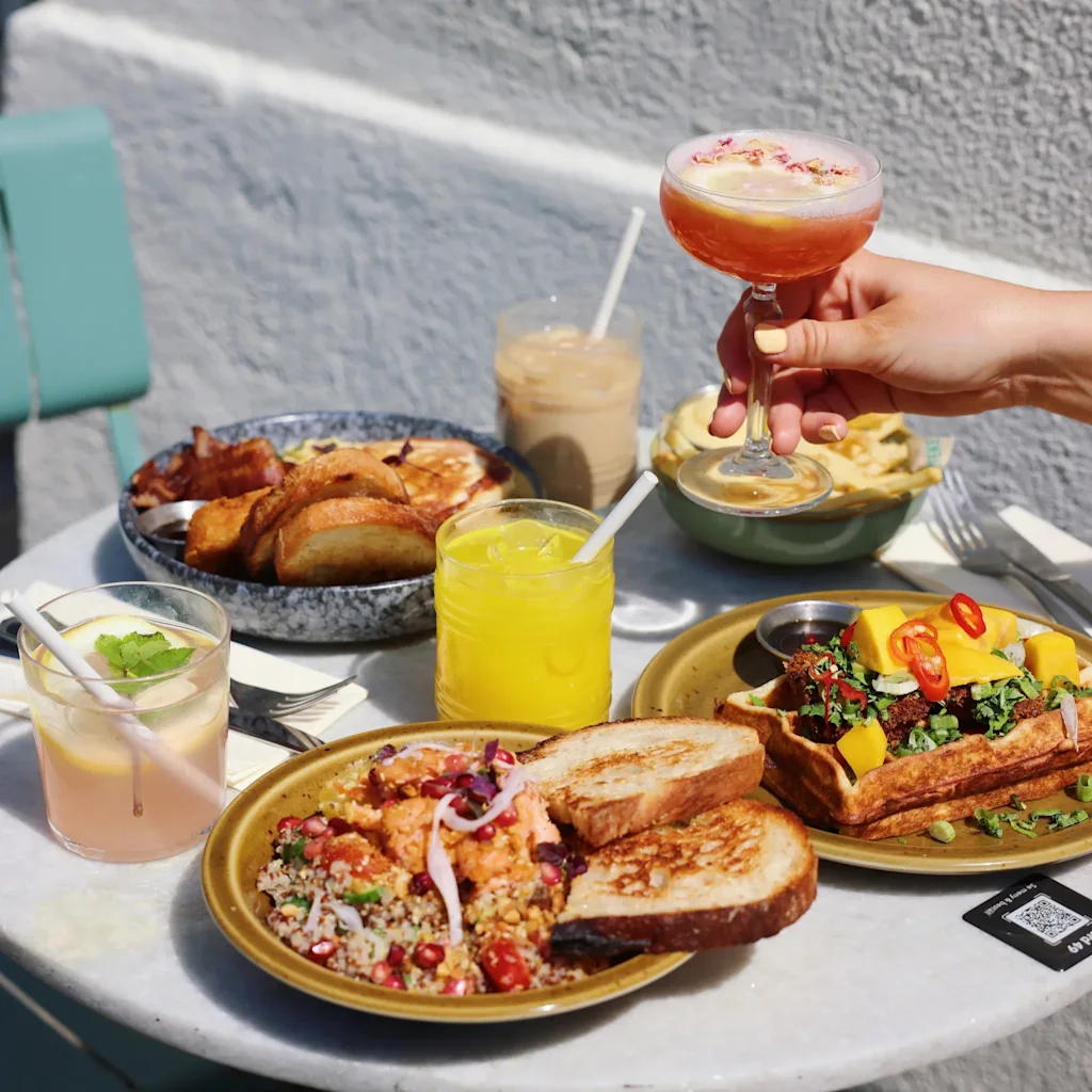 En livlig utomhusbrunch med olika rätter och drycker, inklusive en hand som håller en cocktail. Skanna för meny och lojalitet.