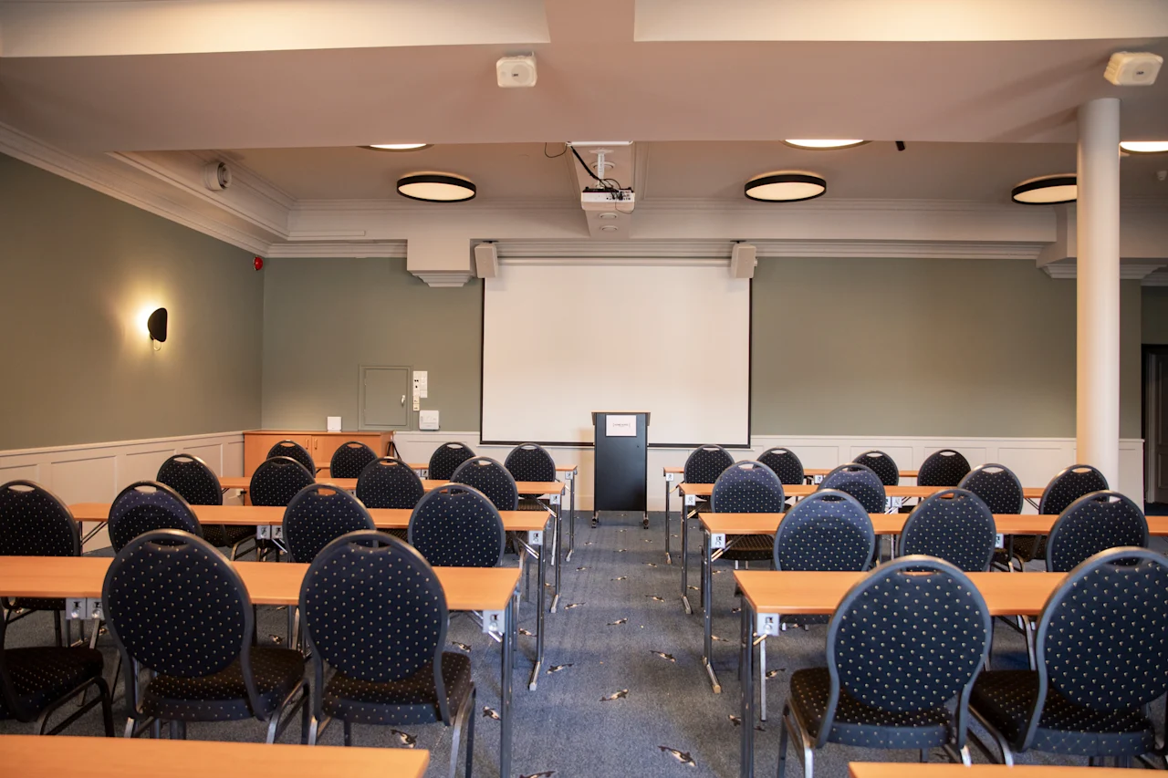 Et veloplyst konferencelokale med rækker af borde og stole vendt mod en stor projektorskærm og et podium.