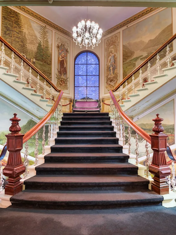 A grand, carpeted staircase with ornate railings leading up to a stained-glass window and murals on the walls.