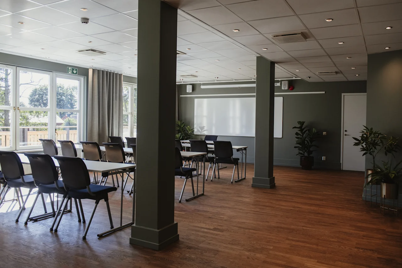 Ett ljust och inbjudande konferensrum med bord och stolar uppställda för ett möte, en whiteboard och stora fönster. En utgångsskylt är synlig.