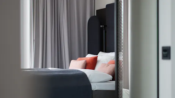 Et koselig hotellrom med en pent oppredd seng, mørk sengegavl, myke puter og elegante grå gardiner.
