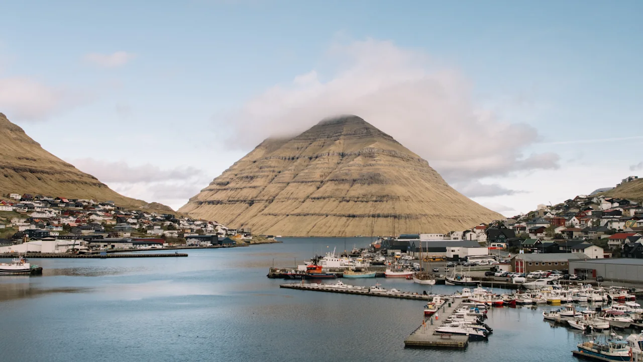 En malerisk havn i en færøsk landsby med en markant, pyramideformet bjergtop i baggrunden under en blå himmel.