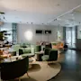 Reception område med grønne stole omkring borde, ved siden af en moderne trædisk med computerskærme. Væggene har indrammet kunst i et lyst, rumligt miljø.