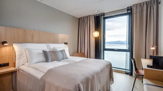 Et hyggeligt hotelværelse med en redt seng, moderne lampe, skrivebord og et stort vindue med udsigt over havet og fjerne bjerge.