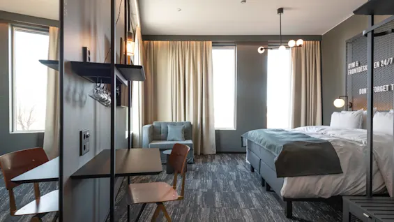A modern hotel room features a bed with a green blanket, a small desk, and a chair. Soft lighting and large windows create a welcoming atmosphere. Text: "GYM & FRONTDESK EN 24/7 DON'T FORGET TO."