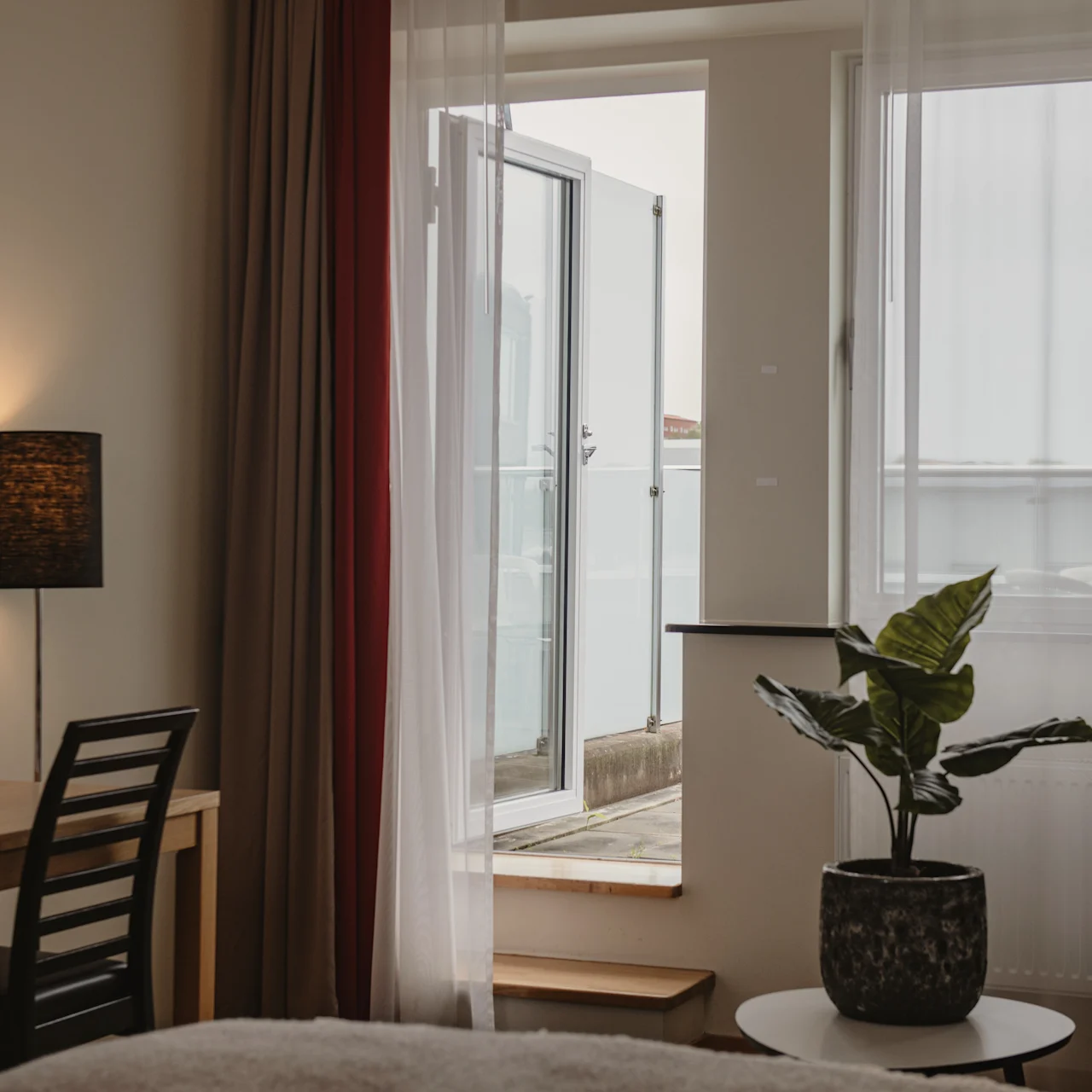 A chair stands next to a lamp and table by a doorway. Sheer curtains frame an open door leading to a balcony. A plant sits on a round table nearby.